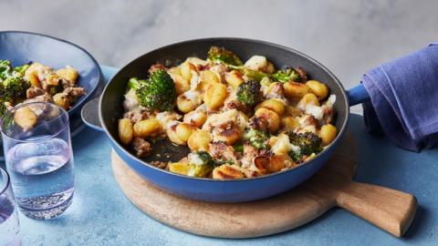 A blue pan sitting on a chopping board. Inside are green vegetables, golden gnocchi and crispy sausage