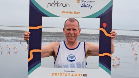 A runner in a white vest is holding up a cardboard photo frame that shows he is in Walvis Bay in Namibia. He is standing in front of a tidal lagoon and there are dozens of pink flamingos on the sand behind him