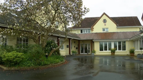 A yellow hospice building with a gabled roof on a rainy day. A tree and shrubs in front of it and a wet concrete driveway.