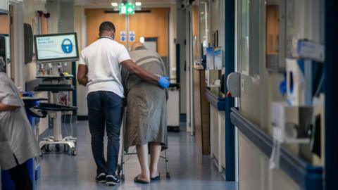 A male nurse supports a male patient as they walk down a hospital corridor, surrounded by hospital equipment