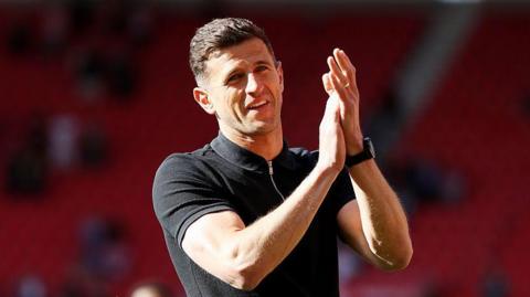 Portsmouth boss John Mousinho wearing a black short sleeved top applauding supporters after a win at Middlesbrough