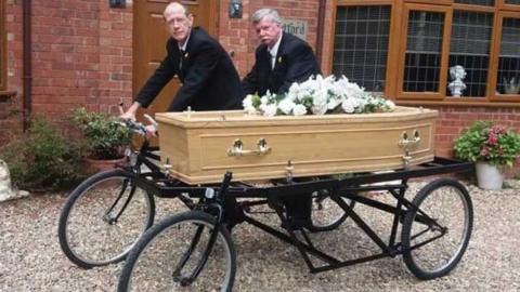 A two-seater tandem bicycle with a coffin attached to the side. Two men are perched on the bicycle and wearing black suits. The coffin is made of pale wood and has white flowers on it.