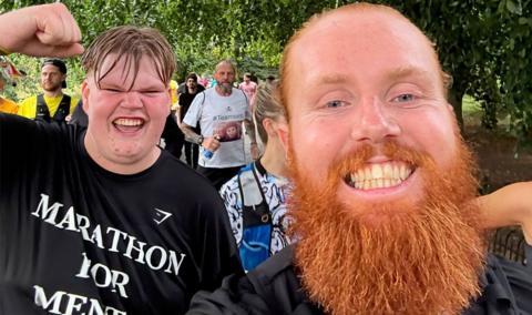 Russ Cook - The Hardest Geezer - and Marcus - The Hull Boy - in the foreground smiling, Marcus has his fist in the air celebrating. They are both wearing black t-shirts. Russ Cook has ginger hair tied back and a ginger beard. Behind them are around 10 runners and trees can be seen surrounding the group.