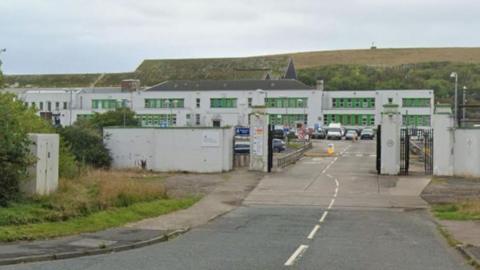 A street view of the Chapel Bank Engineering building in Workington. It's a large white building with green windows. There's a road and car park in front of the building and a large hill behind it.