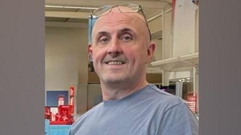 Andrew Blundell is smiling at the camera, he is wearing a blue shirt and glasses on his head and his holding his hand to his chest.