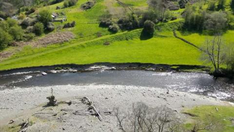 A drone shot of the River Elwy, there is a completely dry grey riverbed next to the river while is now flowing into a nearby green field. In this birds eye view shot there also trees and a couple small buildings. 