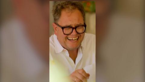 A man, Alan Carr, mid laughter. He wears black glasses and a white shirt.