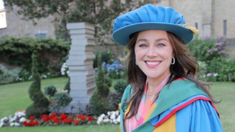 Jill Halfpenny smiles as she looks directly into the camera. She has long dark hair and is wearing a blue cap and gown. She is standing in a garden area.