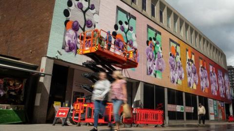 A colourful flowery mural is in the process of being painted on the side of a brick building. A stair lift is raising people up who are working on the mural. There is a street below the mural with people walking past.
