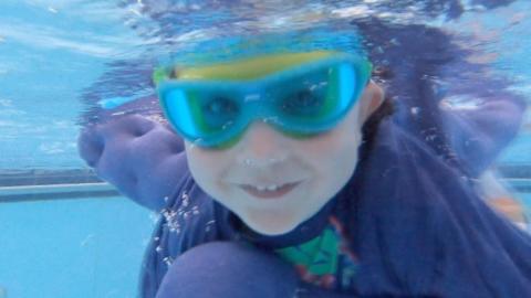 A toddler is underwater in a swimming pool smiling. They are wearing a pair of swimming goggles and a cape is billowing behind them.