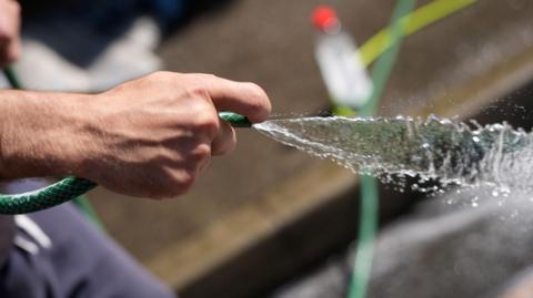 A male hand holds the end of a hosepipe as water gushes out.