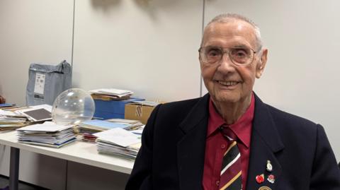 An elderly man with short white hair, large wire-framed glasses and a fairly narrow face smiles at the camera. He wears a red shirt, red, black, white and yellow tie, and black blazer with various poppy and remembrance badges. Behind him on a table are many stacks of cards in envelopes.