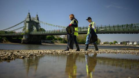 Two volunteers can be seen participating in a clean-up of a Thames riverbank. They are stood on a beach under a green bridge wearing yellow hi-vis vests putting littler into black bin bags using litter pickers.