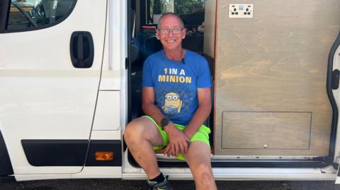 Mark Ferbrache pictured wearing a blue Minions-themed T-shirt, bright green shorts and hiking shoes sitting on the open side doorway of a white camper van. The van’s interior is visible, showing wood-panelled storage units, a power outlet and a sleeping area toward the back.