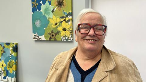 Diabetic Sandra McDermott, who was diagnosed with the disease 20 years ago, is stood in front of floral artworks that are hung on the wall. She has short hair and is wearing a cream jacket, blue and black jumper. Her glasses have a tartan pattern on their frame.