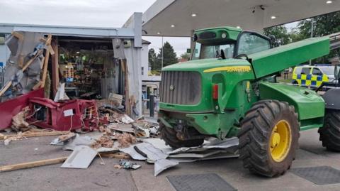 A green telehandler is parked in a garage. It has been used to take a cash machine from the side of a building which has crates outside of it. The wall of the supermarket has been pulled down and there is debris on the floor.