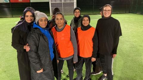 A group of women are looking active. Two people are wearing orange shirts and seem to be in the same team. There is a woman in a blue jumper to the left. Everyone is smiling.