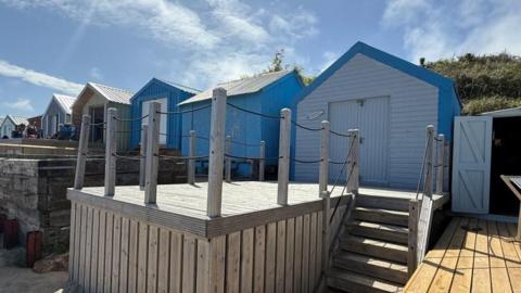 Blue beach hunt with a white double door entrance and wooden decking, and wooden stairs leading down to the beach. The hut is in a row of other beach huts