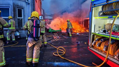 Firefighters in brown suits and yellow helmets on the street holding a hose. There is a fire engine to the right and houses in front. There is a fire on the street which is being tackled by one of the firefighters. To the right of the image is the side of a fire engine with hose reels visible.
