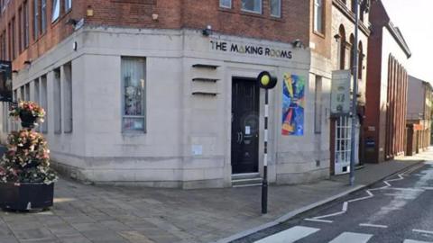 Street view image of the Making Rooms building in Blackburn. The ground floor is white brick and the top floor is red brick. It has long windows and a bright poster next to the black front door and stands next to a pedestrian crossing