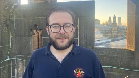 Person is standing outdoors on a high stone structure, likely part of the cathedral tower. They have short brown hair, a brown beard and black framed glasses. Wearing a dark navy polo shirt with a colorful circular emblem on the left chest. The background shows a cityscape with tall buildings and towers under a clear sky. 