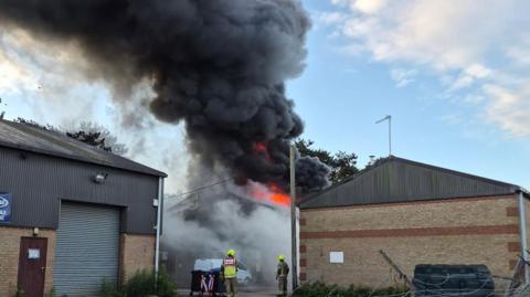 Large plumes of dark smoke billowing out of a brick industrial building. There are some fire crews in the foreground.