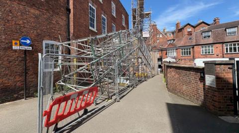 Metal roof spans sit on a road, surrounded by security fencing. Behind it, a fenced-off building is covered in scaffolding.