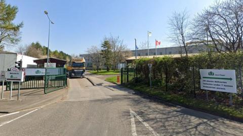 The entrance to a large factory, with a security gate, and a lorry leaving the premises.