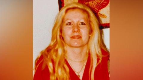A dated image of a blonde woman with dark eye makeup and gold jewellery smiling. The image is portrait and has a blurred background behind it. 