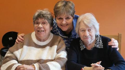 The image shows host Caroline Acreman along with two of her group members.  They are sitting at a dining table and smiling into the camera.