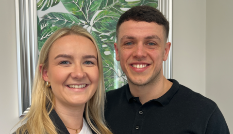 A couple standing shoulder to shoulder in a head and shoulders image. She is on the left and has long blonde hair. He has short dark brown hair. Both have blue eyes and are smiling