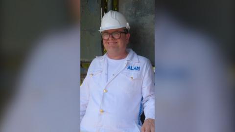 Alan Carr wearing a white helmet and a white boiler suit sitting in a bus stop smiling