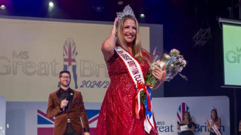 Debbie Hughes is on a stage, there is a sign on the wall behind her that says "Ms Great Britain 2024/2025". She is wearing a sparkly red long dress and a silver crown on her head. She has a sash on that says "Ms Great Britain Classic", and she is holding a bouquet of flowers, and has the other hand on the crown.