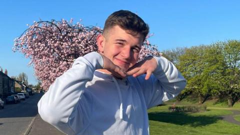 Cole Cooper, a teenage boy, wearing a white hoodie and has his hands placed under his chin in a v-shape as if he is framing his face. He is standing on a pavement next to what looks like a park with grass and a blooming cherry blossom tree behind him.