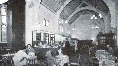 A black and white image from about the 1960s of the former first class waiting room at Carlisle railway station. A number of people are sat at tables covered by gingham table cloths. Two people are standing at the bar on the left. The building has vaulted ceilings painted in white, with some of the wooden structure visible. Large pendant lights hang from the high ceiling.