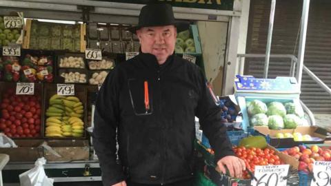 Greengrocer Ben Cooney stands in front of his fruit stall. He is wearing a black fleece, black trousers and a black hat and smiling.