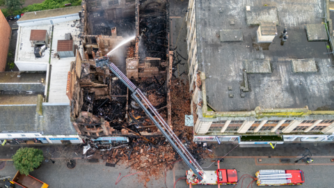 Drone still of the aftermath of the fire, showing a burnt-out building. There are fire engines in the street, including a long turntable ladder with a fireman at the top spraying the building with a hose 