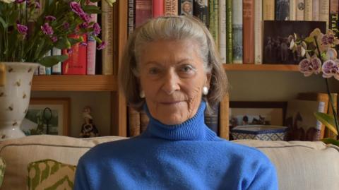 Sue sits on a sofa in her lounge, in front of a bookcase and beside a vase of purple flowers. She is an elderly woman with light-brown grey hair in a hair-band and wears a blue roll-neck sweater.