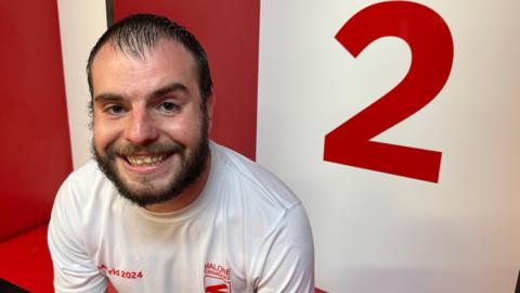 A man in a white t-shirt sitting down with a red number 2 behind him and red locker room wall. He has brown hair, with a brown beard and brown eyes. He is smiling at the camera. 