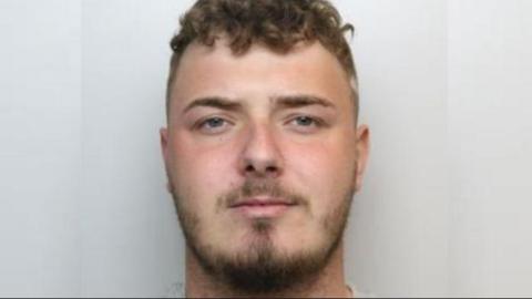 A police custody image of a man with short brown hair and a brown close-trimmed beard.