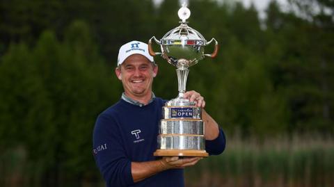Bud Cauley, wearing a dark blue jumper and white cap, smiles and poses with the silver trophy to his left