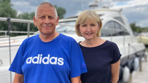 A man in a blue shirt with an Adidas logo and a woman in a dark blue top look towards the camera while standing in front of their boat