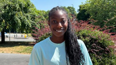 There is a girl in a light blue t-shirt and black hair. Her hair is braided and goes over her left shoulder. She is stood infront of some trees, a grey path to the left of the picture and a roundabout.