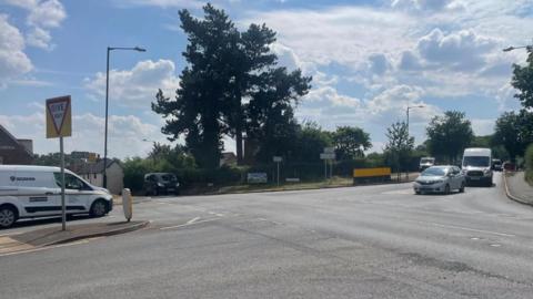 The photo shows a four way crossroads with vans and cars queuing up at each junction to turn left and right. There is a Give Way sign as well as signs pointing to Coventry, Nuneaton, Kingsbury and Meriden. The roads are surrounded by a mix of trees and houses. The sky is blue with some white clouds.