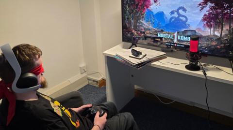 A man wearing a blindfold and headphones is holding a controller and looking at a screen before playing a game called Mortal Combat