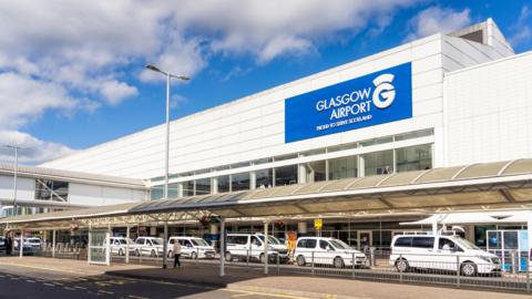 The entrance of Glasgow Airport - a white building with a blue sign - with at least six taxis parked in a rank outside