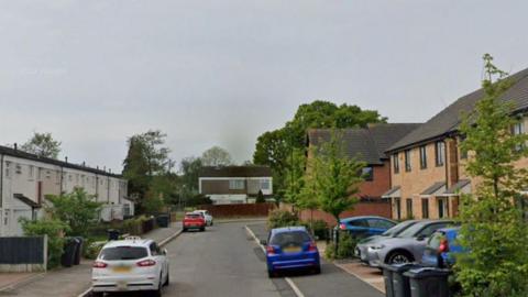 A residential street on a cloudy day. There are two-storey terraced white houses on the left side of the street and two-storey terraced brown brick houses on the right side of the street. There are  a few cars parked on both sides of the street.