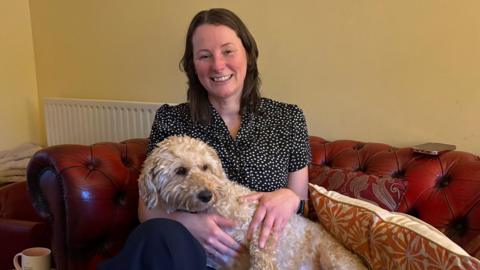 A woman with shoulder length, brown hair is smiling while sitting on a sofa. A white cockerpoo is sitting on her lap. 