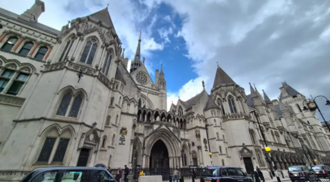 The Royal Courts of Justice building in London. It is a grey concrete building with ornate detailing, turreted roof and archways. There is road traffic in front of the building and blue sky and grey clouds above.