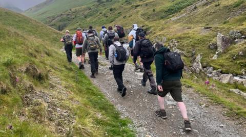 A group of men are climbing a gravel path up a hill all wearing backpacks and walking boots.